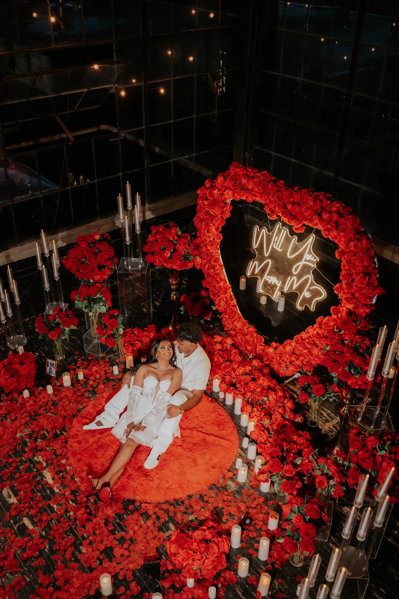 Couple seated among red roses beneath a neon proposal sign